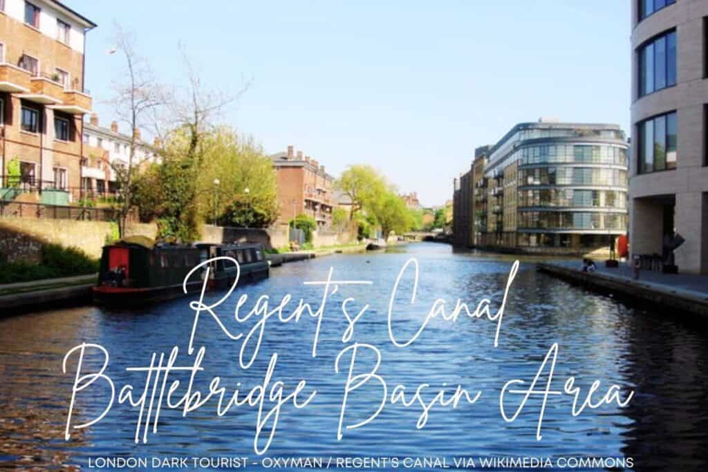 A serene photograph of Regent's Canal at the Battlebridge Basin area in London, featuring a clear blue sky and calm water. This area was not always tranquil - it is where Paula Field's body parts were found floating in carryall bags back in the 1990s. Houseboats are moored along the bank, which is bordered by a mix of residential buildings and modern architecture. Lush green trees add a touch of nature to the urban environment. The script over the image reads 'Regent's Canal Battlebridge Basin Area,' conveying the location's name.